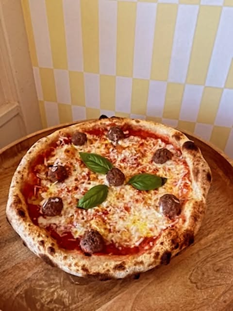 La Polpetta : pizza tomate, fior di latte, polpette, pecorino et basilic, posée sur une planche en bois devant le mur carrelé jaune de la Maison
